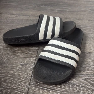 Svarta Adidas sandaler med vita ränder - Klassiska svarta sandaler från Adidas med tre vita ränder över foten. Perfekta att glida runt i på sommaren eller efter träningen. Tillverkade i syntetmaterial och har öppen tå samt platt sula för maximal komfort.