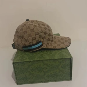 Gucci keps med GG-mönster och blå detalj - Snygg keps från Gucci i beige med klassiskt GG-monogram över hela kepsen. Kepsen har en blå och svart randig detalj på sidan och justerbar rem baktill. Tillverkad i bomullsmix med mörkbrun insida. Perfekt för dig som vill sticka ut med lyxig streetstyle.