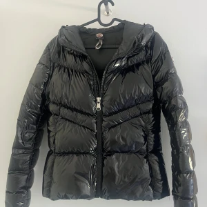 Svart glansig pufferjacka från Colmar - Säljer en svart, glansig pufferjacka från Colmar med huva och dragkedja framtill. Jackan har quiltad design och lång ärm, perfekt för kalla dagar. Snygg och trendig look som passar till många outfits.