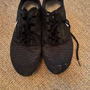 Svarta Nike sneakers med vit sula - Snygga svarta Nike sneakers med vit sula och klassisk vit swoosh på sidan. Ovandelen är i mesh med diskret rutmönster och skorna har snörning. Perfekta för träning eller en sportig vardagslook.