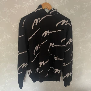 Svart hoodie med vit textprint - Svart hoodie med vit, handskriftsliknande textprint över hela tröjan. Klassisk känguruficka framtill och huva med dragsko. Perfekt för dig som gillar streetstyle och vill sticka ut lite extra. Mjuk och skön, passar till jeans eller joggers.