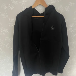 Svart hoodie från Icono Couture, S - Svart hoodie från Icono Couture i slim fit med dragkedja framtill och huva. Snyggt tryck med logga på bröstet och texten 'ICONO COUTURE' i glansigt material längs båda ärmarna. Perfekt för en stilren och modern look.