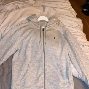 Grå hoodie från Polo Ralph Lauren - Snygg ljusgrå hoodie från Polo Ralph Lauren med dragkedja och huva. Klassisk logga broderad på bröstet och praktiska fickor framtill. Perfekt för chill dagar eller när du vill ha en clean och stilren look.