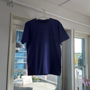 Blå t-shirt från NICCE - Snygg blå t-shirt från NICCE med diskret logga i samma färg på bröstet. Klassisk rund halsringning och korta ärmar. Perfekt basic-plagg med stilren vibe, passar till jeans eller shorts. Skön och mjuk känsla.