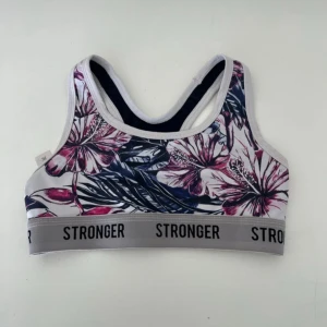 Blommig sporttopp från Stronger XS - Sporttopp från Stronger i storlek XS med blommigt mönster i rosa, lila och mörkblå toner. Elastiskt grått band med logga runt om. Perfekt för träning och har en sportig, ärmlös design med brottarrygg. Materialet är mjukt och följsamt syntet.