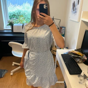 Blommig ljusblå klänning  - Supersöt ljusblå kortklänning från Vero Moda med vitt blommönster, volang och knyt i midjan. Klänningen har korta ärmar och en avslappnad passform, perfekt för varma dagar. Materialet känns lätt och luftigt. Passar även om man brukar ha storlek XS.