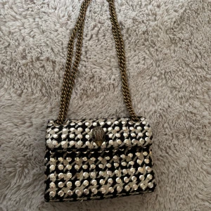 Svart och silver axelväska med kedja - Kurt Geiger axelväska i svart tyg med guldiga paljetter och broderat rutmönster. Superfin till fest eller vardag!! Några paljetter har lossnat från väskan men inget som tar ifrån helheten av väskan enligt mig💕 Går ej att köpa längre, så den är unik🥰 Skriv privat om du vill köpa så fixar vi!!
