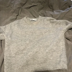 Grå stickad tröja från Zara - Mysig grå stickad tröja från Zara i storlek M. Tröjan har korta ärmar, rund halsringning och en enkel, clean look. Perfekt för lager-på-lager och chill dagar. Materialet känns mjukt och passar bra till både jeans och kjol.