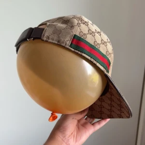 Gucci keps med GG-mönster och röd/grön rand - Säljer en snygg Gucci keps i beige med klassiskt GG-monogram över hela kepsen och ikonisk röd/grön rand på sidan. Justerbar rem baktill i brunt skinn. Kepsen är tillverkad i bomull och har en cool streetstyle-vibe. TVEKA INTE VID FRÅGOR/ PRIS KAN DISKUTERAS