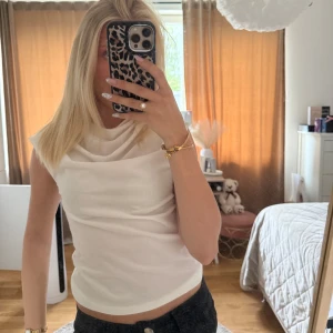 Vit ärmlös draperad topp - Säljer en stilren vit topp med draperad halsringning och ärmlös design. Toppen har en mjuk, lätt figurnära passform och är perfekt för att skapa en clean look. Passar snyggt till både jeans och kjol.