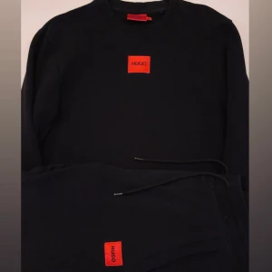 Svart dress från Hugo Boss  - Svarta mjukisbyxor och tröja  från Hugo Boss med röd HUGO-logga framtill. Byxorna har dragsko i midjan och raka ben. Tillverkade i mjukt bomullsmaterial för en chill och snygg look. Pris kan diskuteras