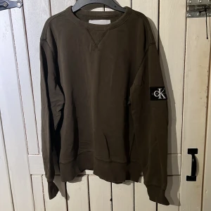 Olivgrön sweatshirt från Calvin Klein - Olivgrön sweatshirt från Calvin Klein Jeans med klassisk rund hals och ribbade muddar. Stor CK-logga på ärmen ger en clean och stilren vibe. Perfekt för dig som gillar enkel men snygg streetstyle.