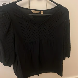 Svart blus med broderade detaljer Culture - Svart blus från Culture i storlek XL med snygga broderade mönster och håldetaljer på både bröst och ärmar. Blusen har v-ringning och trekvartsärmar, vilket ger en chill och luftig vibe. Perfekt för dig som gillar stilrena men ändå unika plagg.