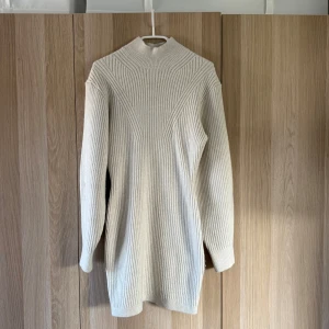 Beige ribbad stickad klänning H&M - Supermysig beige stickad klänning från H&M med ribbad struktur och hög krage. Långärmad och i kort modell, perfekt för lager på lager. Snyggt mönster upptill som ger extra detalj. Passar dig som gillar enkel men trendig stil.