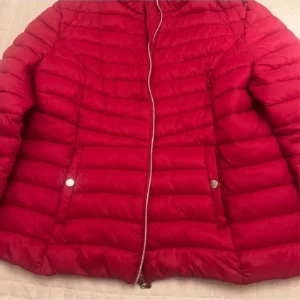 Röd pufferjacka med dragkedja - Snygg röd pufferjacka med horisontella sömmar och silverfärgad dragkedja framtill. Jackan har två fickor med tryckknappar och en klassisk, sportig look. Perfekt för kyliga dagar när du vill sticka ut lite extra.