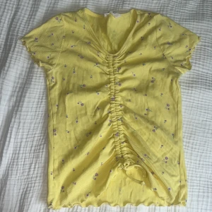 Gul blommig topp med snörning H&M - Superfin gul topp från H&M med små lila blommor och ribbad struktur. Toppen har korta ärmar, v-ringning och snygg snörning framtill som ger en cool rynkad effekt. Perfekt för vår och sommar när du vill ha något färgglatt och bekvämt.