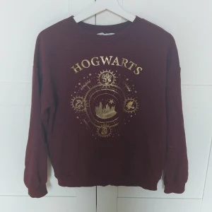 Vinröd Hogwarts-tröja från H&M - Vinröd sweatshirt från H&M med stort Hogwarts-tryck i guld på bröstet. Tröjan har rund hals, långa ärmar och ribbade muddar. Perfekt för Harry Potter-fans som vill sticka ut med magiska detaljer och snygg färg. Det är inget fel på tröjan men den har blivit för liten.