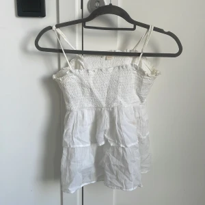 Vit volangtop 🤍 - Supersöt vit topp från Gina Tricot med smala axelband och smockad överdel. Toppen har flera lager volanger som ger en luftig och somrig känsla. Perfekt till shorts eller kjol för en chill look. Denna har lite fläckar på sig!!