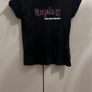 Svart Replay t-shirt med tryck - Svart t-shirt från Replay med coolt tryck i lila och vitt framtill. Klassisk passform med korta ärmar och rund hals. Perfekt till jeans eller shorts för en avslappnad look.