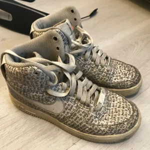 Nike Air Force 1 High Just Do It - Unika Nike Air Force 1 High med heltryckt 'Just do it'-text över hela skon. Beige och vita toner, högt skaft och klassisk snörning. Swoosh-logga på sidan och Nike Air-brodyr på hälen. Ovandel i skinn och textil för en cool och urban vibe.