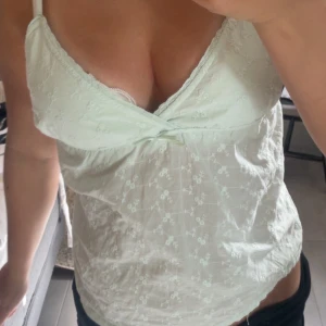 Ljusgrön blommig linnetopp - Supersöt ljusgrön linnetopp med tunna axelband och små broderade blommor över hela tyget. Toppen har v-ringning och en liten rosett framtill. Perfekt för varma dagar och ger en fräsch och romantisk vibe.