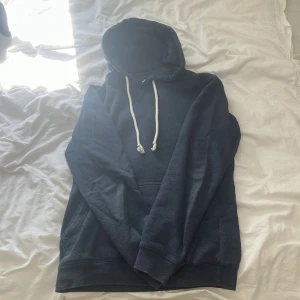 Mörkblå hoodie med vita snören - En stilren mörkblå hoodie med klassisk känguruficka och vita dragsnören i huvan. Tröjan har lång ärm och en relaxed passform, perfekt för chill dagar. Materialet känns mjukt och skönt mot huden, och den funkar lika bra till jeans som till mjukisbyxor.