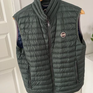 Mörkgrön dunväst från Colmar - Snygg mörkgrön dunväst från Colmar med quiltad design och hög krage. Västen har dragkedja framtill, två fickor med dragkedja och en diskret logga på bröstet. Perfekt att slänga över en hoodie eller tröja för extra värme.