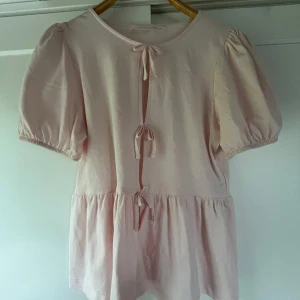 Ljusrosa blus med knyt från Gina Tricot - Supersöt ljusrosa blus från Gina Tricot med puffiga korta ärmar och tre dekorativa knyt framtill. Blusen har en volang nedtill💕