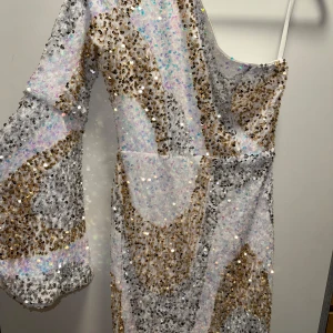 Glittrig långklänning med paljetter - En superglittrig långklänning med swirlmönster i silver, guld och vitt. Klänningen har en ärm och en axelbandslös sida, vilket ger en cool oneshoulder-look. Täckande med massor av paljetter som reflekterar ljuset. Perfekt för dig som vill sticka ut.