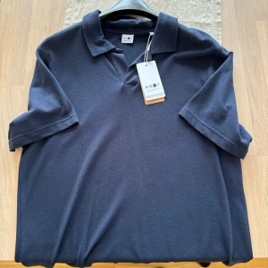  NN07 - Snygg mörkblå pikétröja från NN07 med klassisk krage och korta ärmar. Tröjan är tillverkad i ett mjukt och skönt material som känns lyxigt mot huden. Helt ny aldrig använd nypris 1299kr
