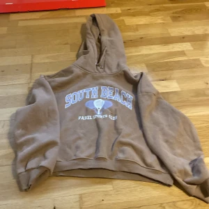 Beige hoodie South Beach 157 M - Säljer en beige hoodie från 157 i storlek M med trycket 'South Beach Padel Sports Club' i blått och vitt framtill. Tröjan har huva och ribbade muddar vid ärmslut och nederkant. Perfekt för en avslappnad och sportig stil.