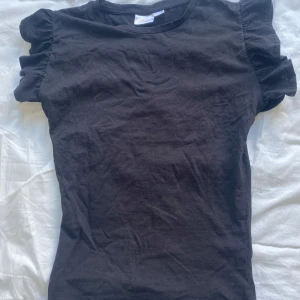 Svart topp med volangärmar Gina Tricot - Svart topp från Gina Tricot med korta volangärmar som ger en söt och trendig look. Tillverkad i mjuk bomull och har en klassisk rund halsringning. Perfekt att styla med jeans eller kjol för en enkel men snygg outfit.