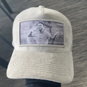 Beige mockakeps med fototryck - Snygg beige keps i mockaimitation med mesh på sidorna och baksidan för extra ventilation. Framtill finns ett svartvitt fototryck av en firande fotbollsspelare. Kepsen har böjd skärm och klassisk snapback-stil. Perfekt för dig som vill sticka ut med en unik accessoar.