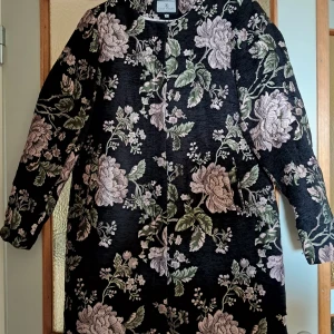Blommig svart kappa från Flash Woman - Snygg svart kappa med stort blommönster i lila och grönt från Flash Woman. Jackan har långa ärmar, dold knäppning med tryckknappar och rak passform. Perfekt för dig som vill sticka ut med en unik och trendig look. - Fint skick :)