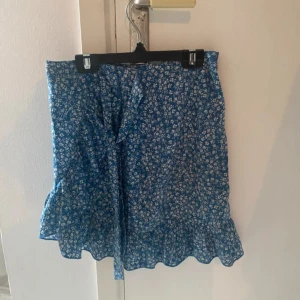Blå blommig volangkjol från SHEIN - Supersöt blå kortkjol från SHEIN med vitt blommönster och volangkant nertill. Kjolen har omlottkänsla och stängs med dragkedja i sidan 