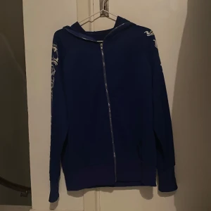 Blå hoodie med tryck från SHEIN - Blå hoodie från SHEIN med hel dragkedja och huva. Ärmarna har vita tryck i gotisk stil längs hela längden. Tröjan har ribbade muddar och fickor framtill. 