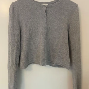 Grå croppad kofta  - Grå croppad kofta från H&M Basic med rund halsringning och knappar framtill. Långärmad och i mjuk stickad bomullsblandning, perfekt att slänga över ett linne eller t-shirt. Enkel och stilren design som funkar till allt.