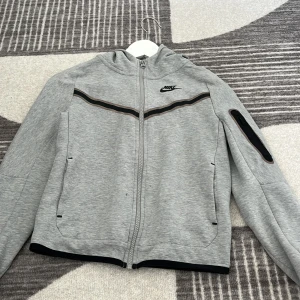 Grå zip-hoodie från Nike med svarta detaljer - Grå zip-hoodie från Nike med svarta detaljer och logga på bröstet. Jackan har hel dragkedja, huva och en cool ficka med dragkedja på ärmen. Perfekt för dig som gillar sportig och avslappnad stil.