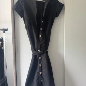 Svart Trendyol kortklänning med knappar - Svart kortklänning från Trendyol med snygga guldiga knappar framtill och tillhörande bälte i midjan. Klänningen har korta ärmar och markerad midja för en feminin siluett. Perfekt för dig som gillar stilrena och enkla plagg.