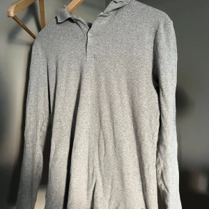 Grå långärmad pikétröja - Säljer en stilren grå långärmad pikétröja i mjuk bomull. Tröjan har klassisk krage och knäppning framtill, perfekt för en clean och avslappnad look. Passar dig som gillar enkel och tidlös stil. Har fett fläckar som man knappt ser.