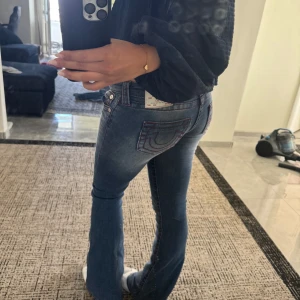 Blå bootcut jeans från True Religion - Säljer ett par blå bootcut jeans från True Religion med ikoniska bakfickor och röda sömmar. Jeansen har låg midja och är tillverkade i klassiskt denimtyg med snygg tvätt och lätt slitning. Perfekta för dig som gillar en retro vibe och vill sticka ut. Storlek 25 (xs-s) helt nya och aldrig använda. Köpta feån nelly
