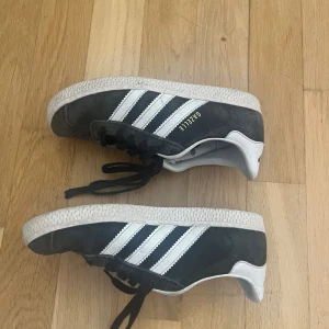 Adidas Gazelle svarta sneakers - Klassiska Adidas Gazelle sneakers i svart mocka med vita detaljer och de ikoniska tre ränderna på sidan. Skorna har snörning och en platt vit sula. Perfekta för en sportig och stilren look.