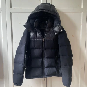 Ralph Lauren Jacka - Snygg svart dunjacka från Polo Ralph Lauren med glansiga detaljer upptill och stor huva. Jackan har quiltad design, dragkedja framtill och är perfekt för kalla dagar. Klassisk look med en modern twist, passar dig som vill ha både stil och värme.