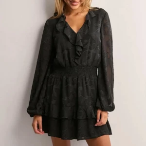 Nelly, Svart kort volangklänning  - Supersöt svart kort klänning från Nelly med v-ringning och volangdetaljer längs halsringningen. Långa ärmar i transparent tyg, smockad midja och dubbla volanger i kjolen. Tunt, lätt material med diskret blommönster i tyget. Köpt för 399 kr och använd endast 1 gång!