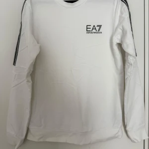 Vit långärmad tröja EA7 Armani - Snygg vit långärmad tröja från EA7 Emporio Armani med svarta ränder och logga på bröstet. 