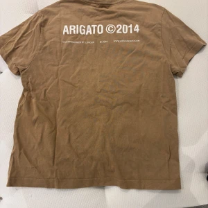 Beige t-shirt från Axel Arigato - Snygg beige t-shirt från Axel Arigato med tryck både fram och bak. Klassisk rund halsringning och korta ärmar. Tillverkad i 100% ekologisk bomull för en skön och avslappnad känsla. Perfekt för dig som gillar stilrena basplagg med en twist.