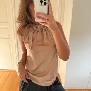 Beige ärmlös linnetopp med knyt - Säljer en beige ärmlös linnetopp med snygg knytning vid axeln och lätt glansigt material. Nyskick 