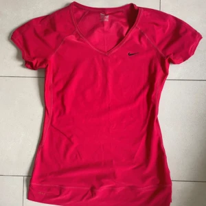 Röd sporttopp från Nike - Nike sporttopp i stark röd färg med v-ringning och korta ärmar. Toppen har en slim passform och är tillverkad i ett lätt, syntetiskt material som andas. Perfekt för träning och aktiva dagar. Liten svart Nike-logga på bröstet för en clean look.
