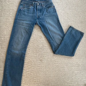 Levi's 501 blå jeans W31 L34 - Klassiska Levi's 501 jeans i mellanblå tvätt med raka ben och knappgylf. Snygg vintagekänsla och ikonisk läderpatch bak i midjan. Jeansen har fem fickor och orange sömmar. Perfekta för dig som gillar tidlös denimstil.