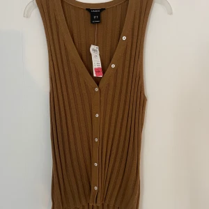 Ribbad brun väst från Lindex - Säljer en ribbad brun väst från Lindex med v-ringning och knappar framtill. Västen är ärmlös och har en snygg, vertikal ribbstruktur som ger en modern look. Perfekt att styla över en t-shirt eller skjorta för en trendig layering. HELT oanvänd med prislapp fortfarande på